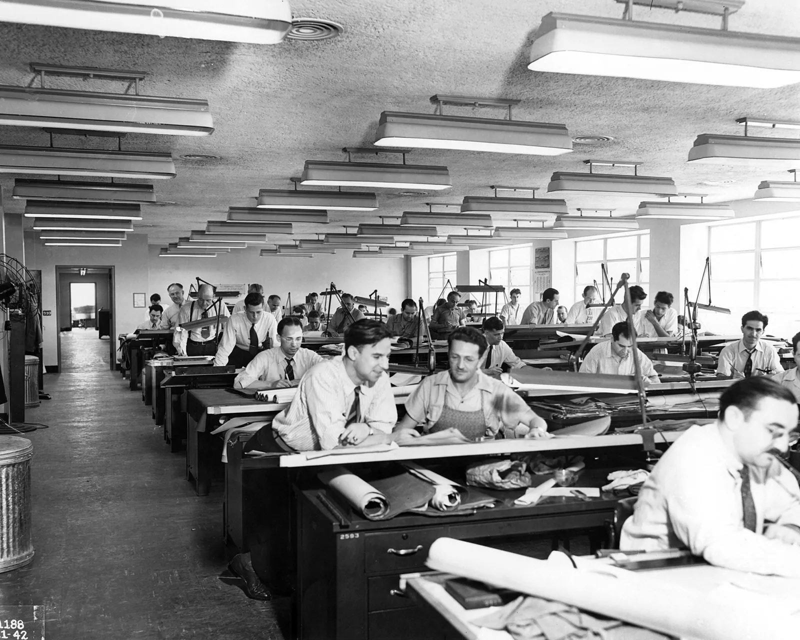 Rows of engineers at drafting tables in a long office lit by ceiling lamps