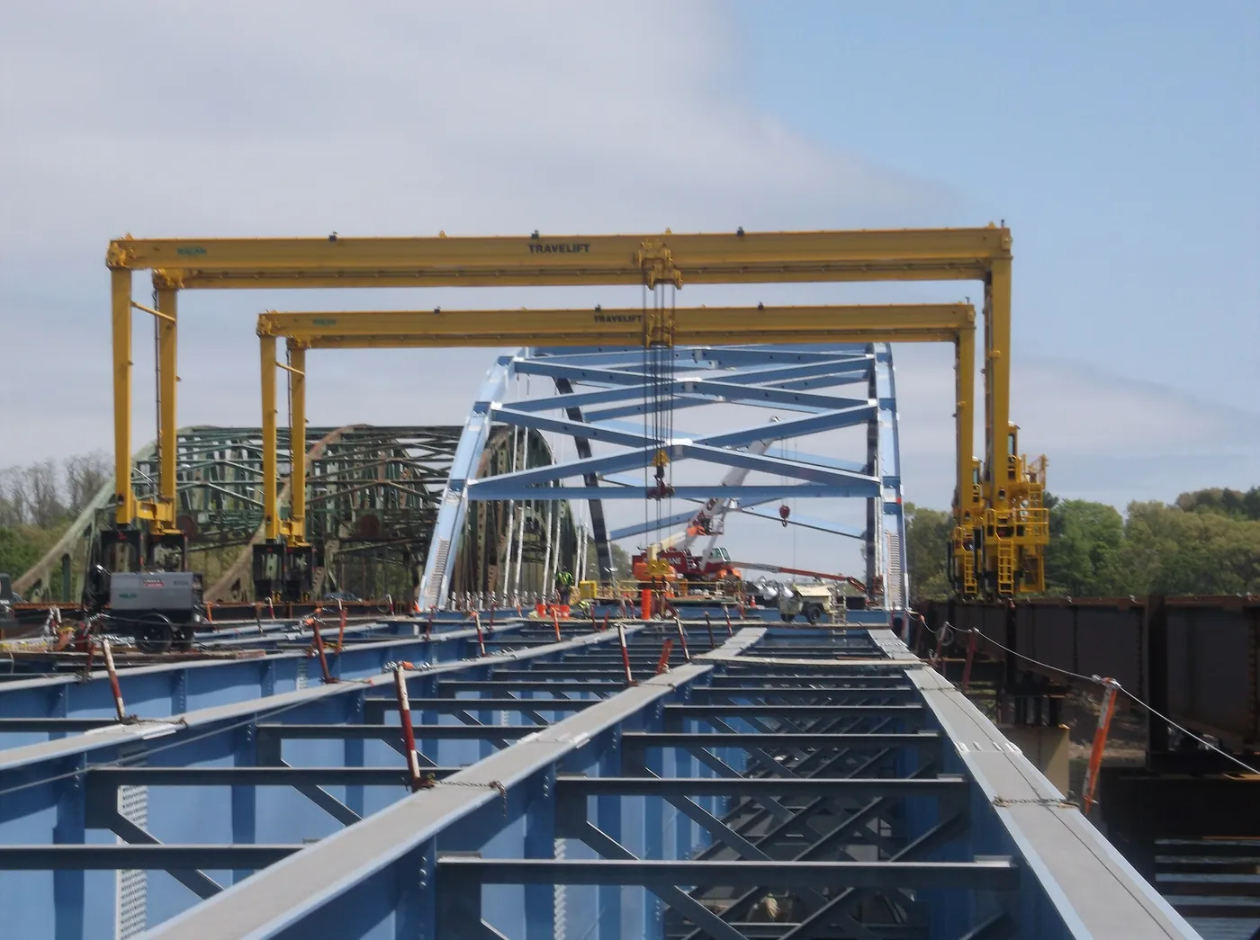 Steel girders being placed on a bridge deck between two yellow gantry cranes